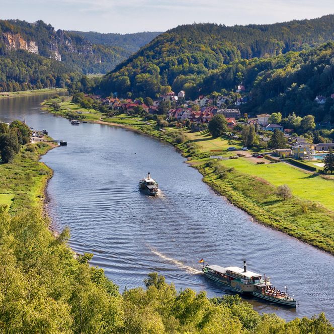 Blick auf einen Fluss, umgeben von grünen Hügeln und Bäumen, mit Booten auf dem Wasser.