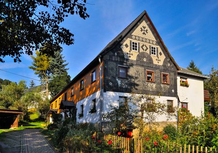 Fachwerkhaus mit bunten Fensterläden und Garten, umgeben von Bäumen und blauem Himmel.