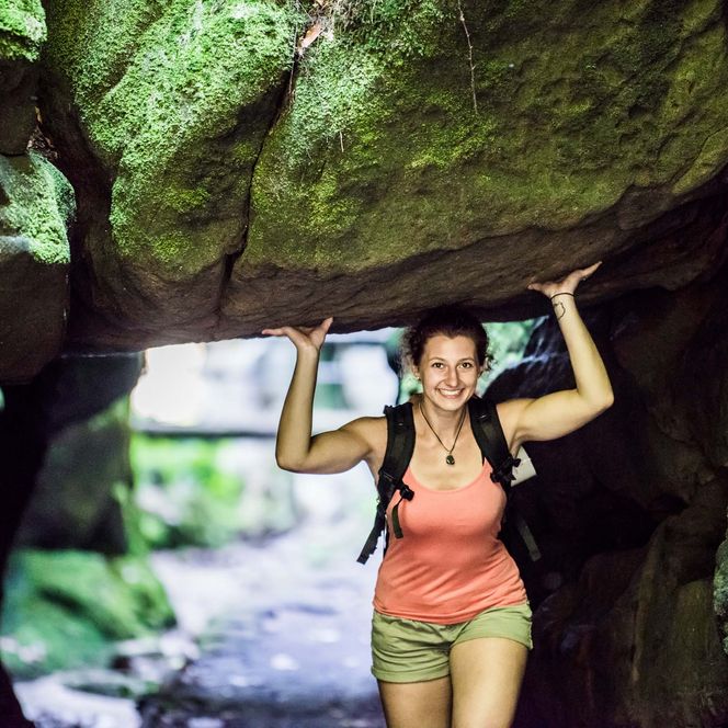 Frau mit Rucksack "hebt" lachend einen großen Felsblock des Uttewalder Felsentor über ihrem Kopf in einer Höhle.