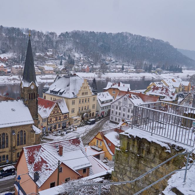 Schneebedeckte Stadt mit Häusern und Kirchturm, umgeben von Hügeln und einem Fluss im Hintergrund.