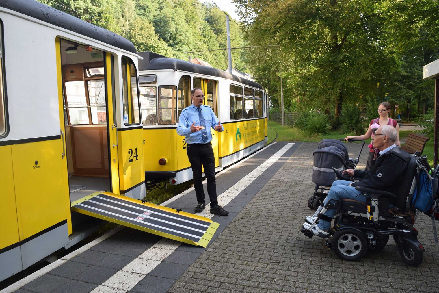 Mann in Uniform steht neben einem Zug mit ausklappbarer Rampe, während eine Frau mit Rollstuhl wartet.