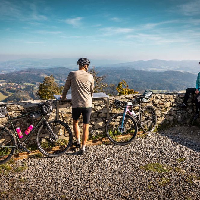 Zwei Radfahrer stehen an einer Aussichtspunkt mit Blick auf eine Berglandschaft, Fahrräder stehen neben ihnen.