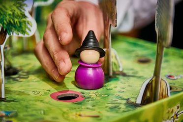 A hand reaches for a purple game piece wearing a black hat on a game board depicting a green landscape.