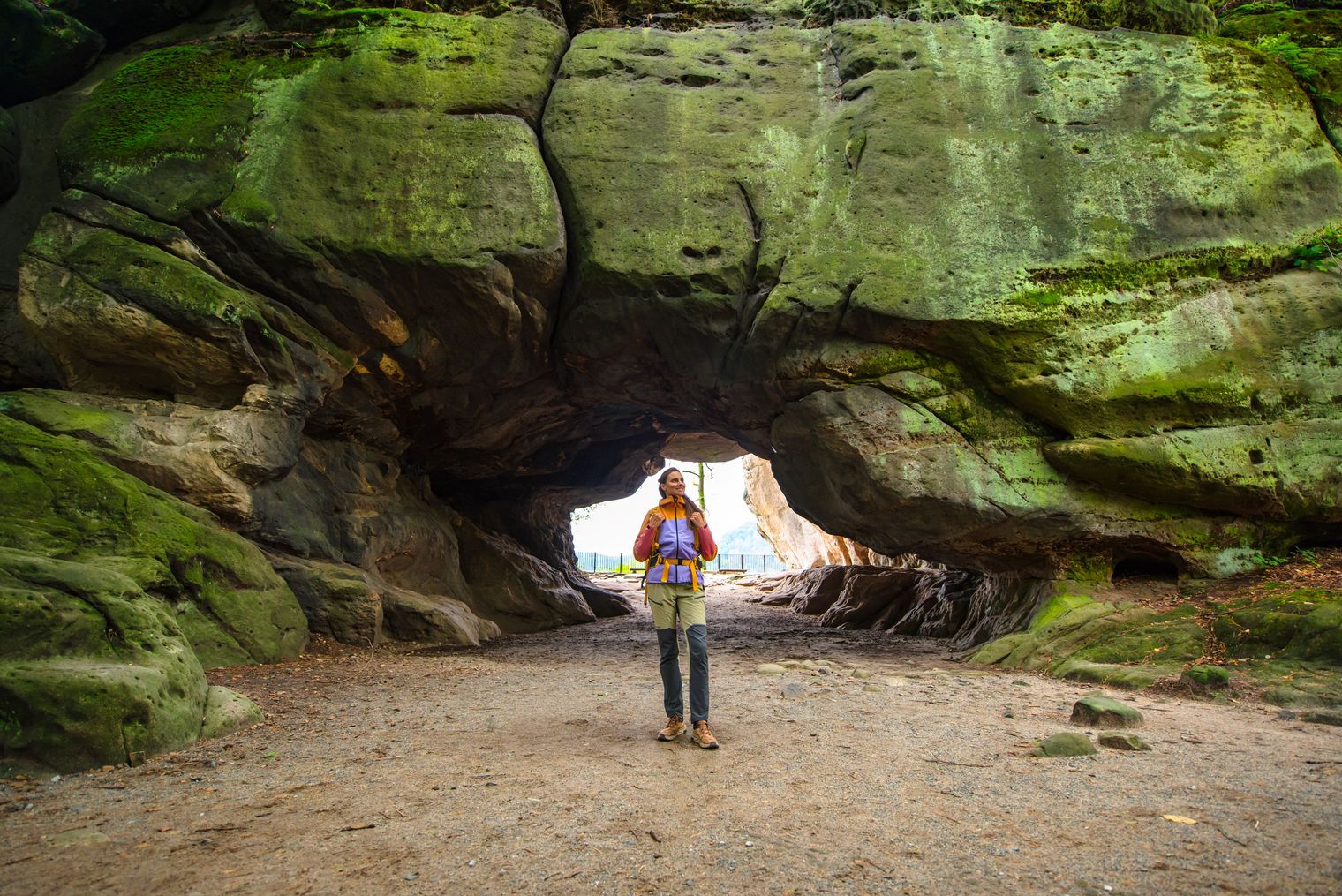 Person mit Rucksack steht vor einer großen, moosbedeckten Felsöffnung (Kuhstall) in einer Höhle.