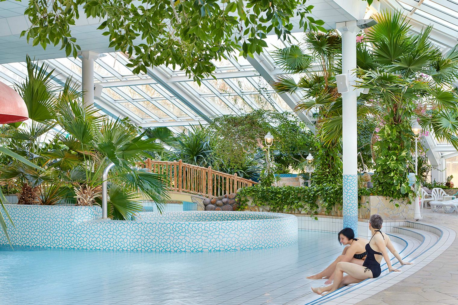 Zwei Personen sitzen am Rand eines Schwimmbeckens in einer tropischen Umgebung mit Pflanzen.