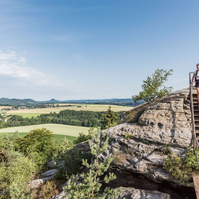 Wanderer spaziert auf einer Felsformation und blickt auf eine weite Landschaft mit Feldern.
