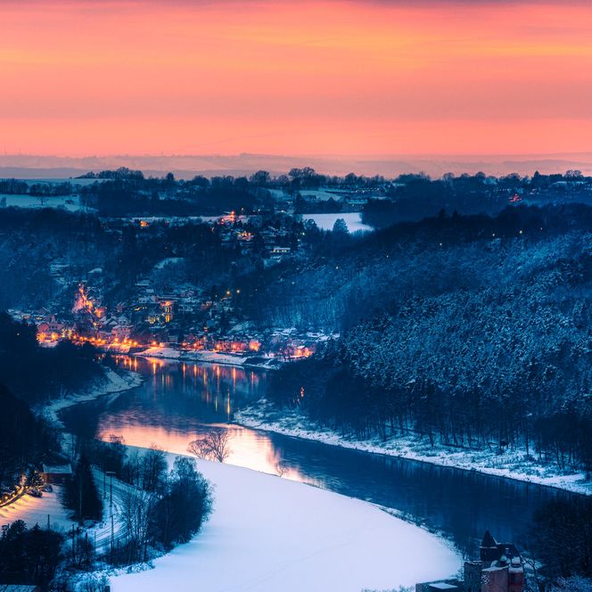 Blick auf einen Fluss, umgeben von schneebedeckten Bäumen und einem Dorf, bei Sonnenuntergang.