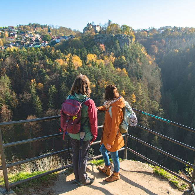 Zwei Personen stehen an einem Aussichtspunkt und blicken auf die herbstliche Landschaft des Polenztals mit Wäldern und Hügeln und der Burg Hohnstein im Hintergrund.
