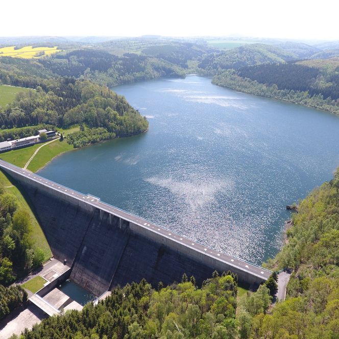 Luftaufnahme eines Staudamms mit Wasserreservoir und umgebender grüner Landschaft.