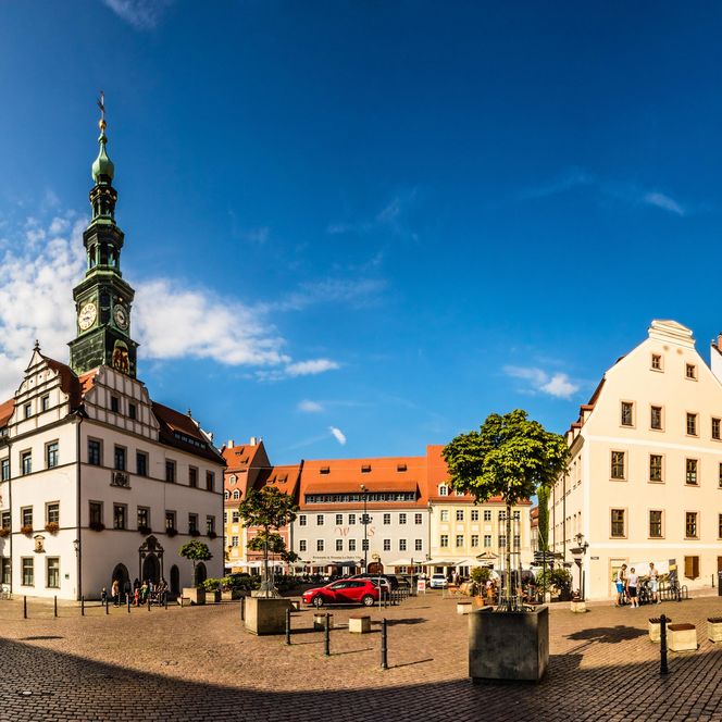 Panoramaansicht eines historischen Platzes mit mehreren Gebäuden und einem Kirchturm unter blauem Himmel.