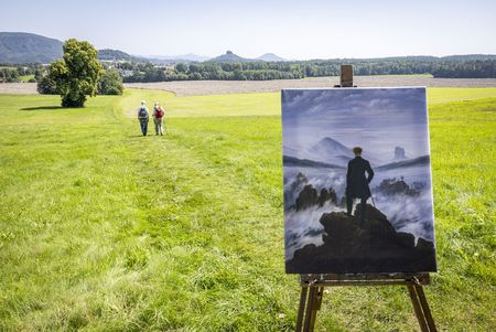 Zwei Personen gehen auf einem Feld, im Vordergrund steht ein Gemälde auf einer Staffelei.