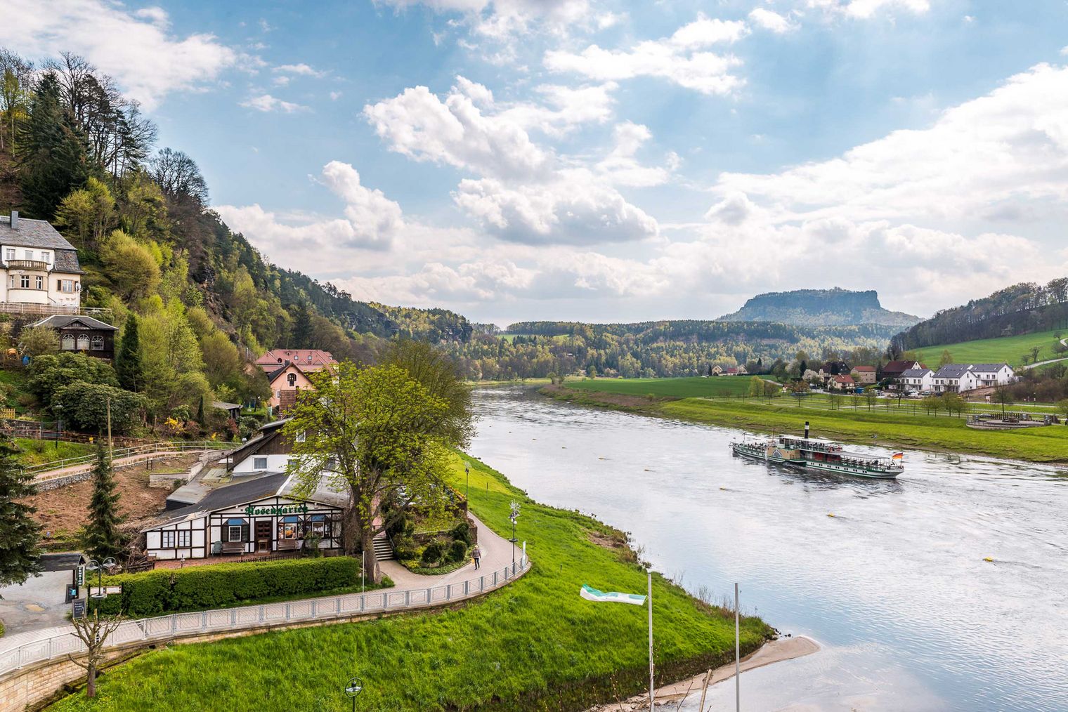 Flusslandschaft der Elbe mit grünen Ufern, einem Boot der Dampfschifffahrt auf dem Wasser und bewaldeten Hügeln und dem markanten Tafelberg Lilienstein im Hintergrund.