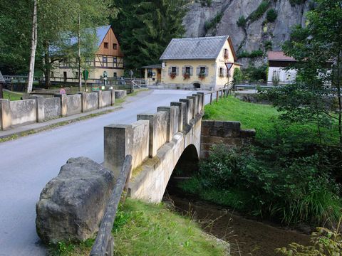 Historisches Mühlengebäude in ruhiger Waldlandschaft bei klarem Himmel, im Vordergrund eine Steinbrücke.