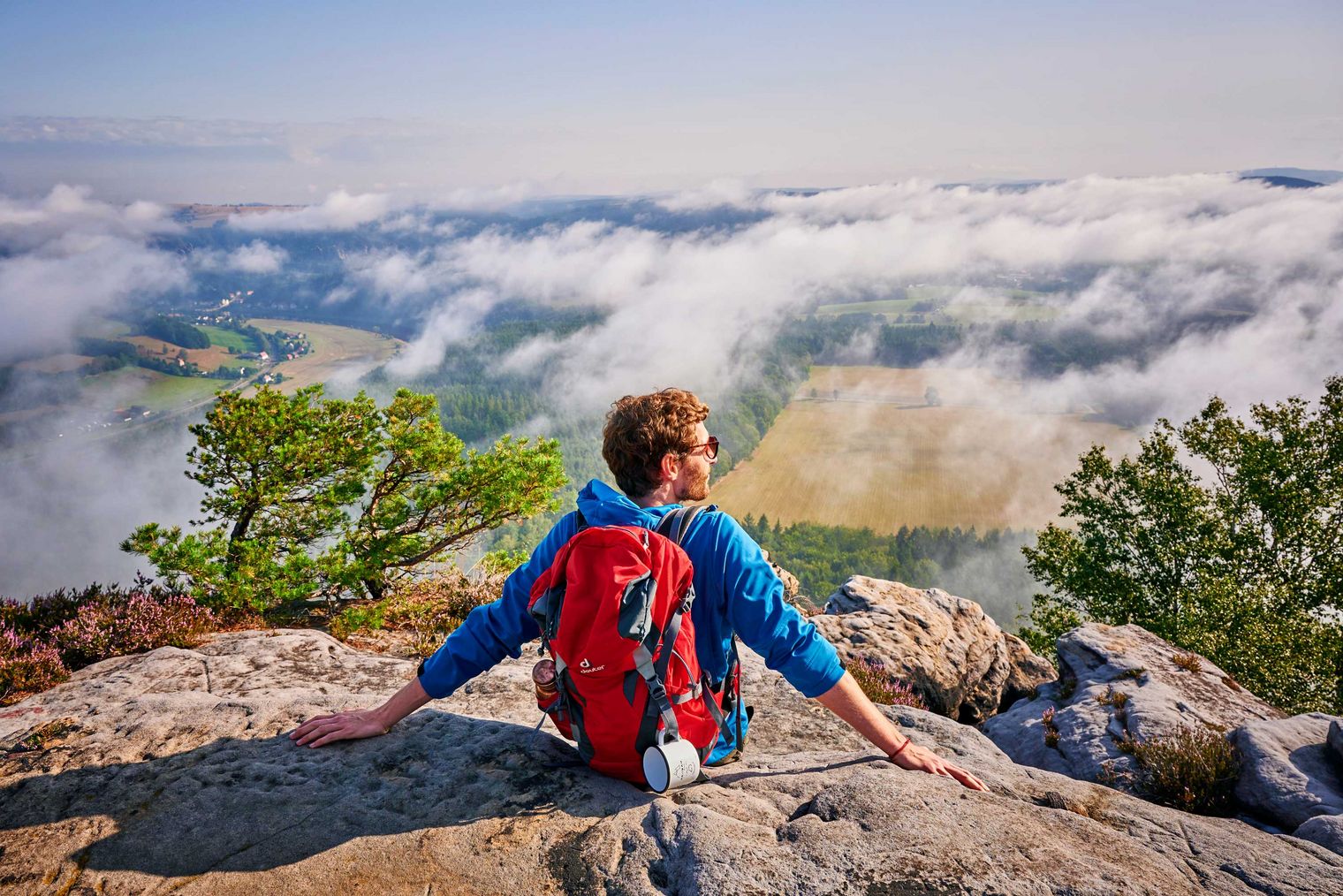 Person mit Rucksack sitzt auf einem Sandstein-Felsen und blickt auf eine nebelverhangene Landschaft.