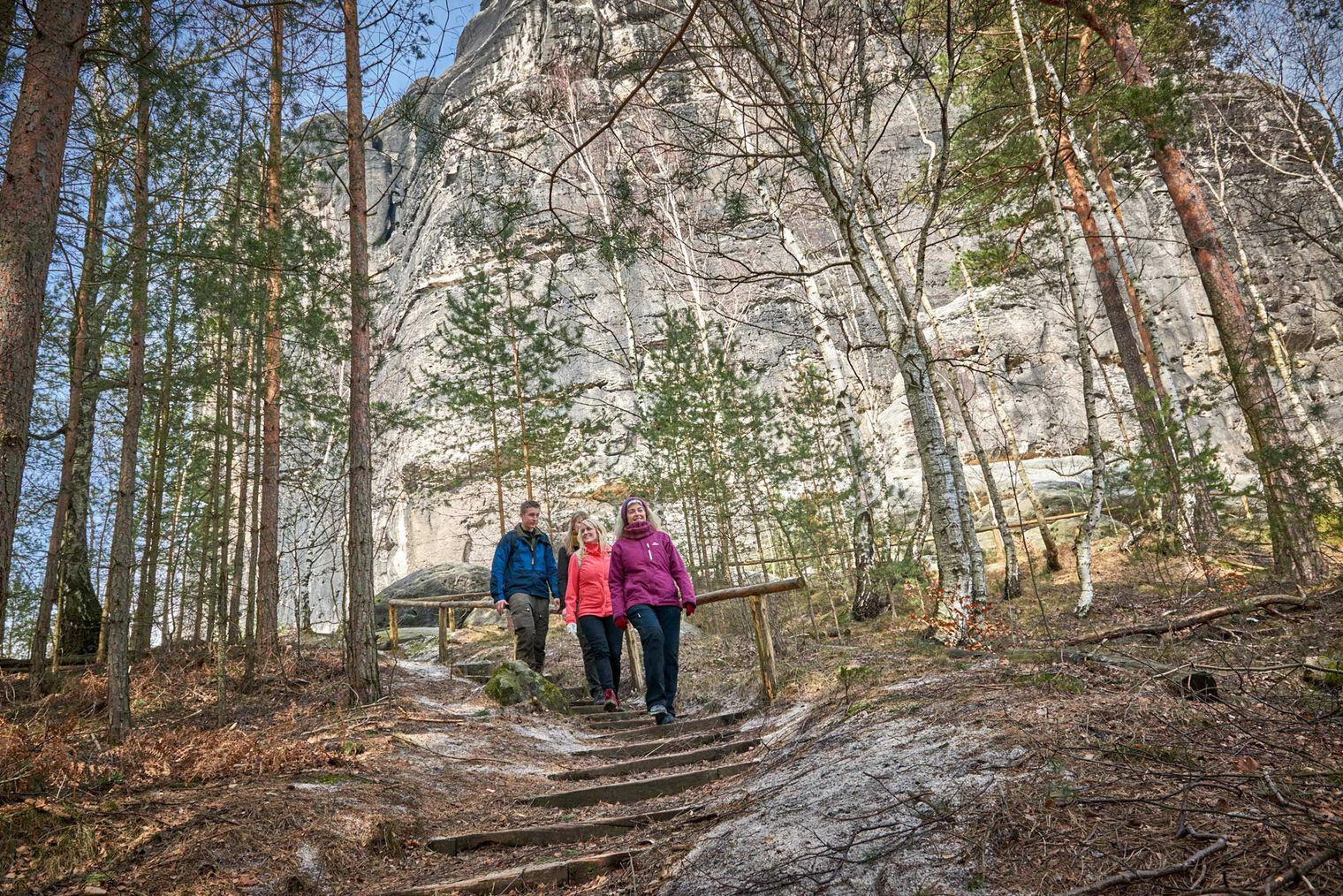Zwei Personen wandern auf einem schmalen Weg durch einen Wald mit Felsen im Hintergrund.
