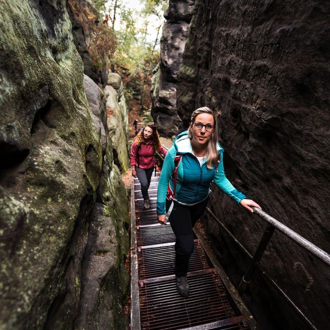 Zwei Personen steigen eine steile Treppe in einer schmalen Schlucht hinauf, umgeben von Felsen.