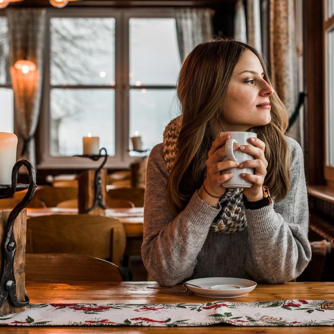 Frau mit einer Tasse in der Hand, sitzt an einem Tisch in einem gemütlichen, winterlichen Café.
