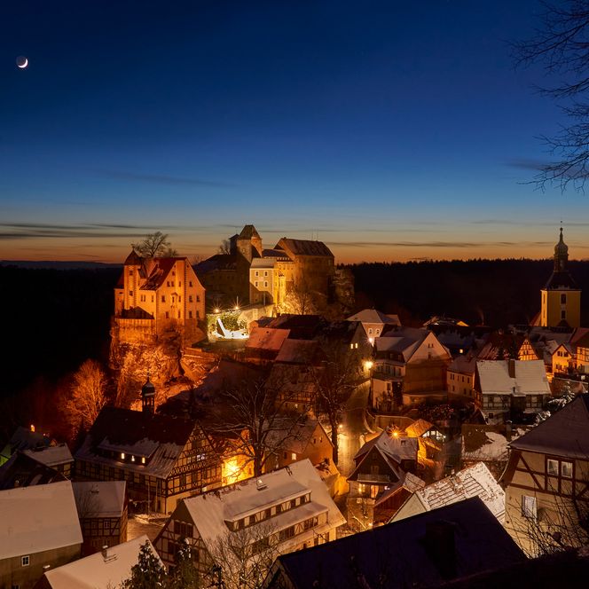 Dämmerungsansicht einer Stadt mit beleuchteten Gebäuden und Felsen im Hintergrund. Ein Stern leuchtet am Himmel.