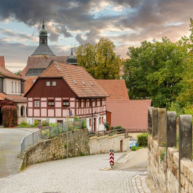 Historisches Fachwerkhaus mit roten Dächern, umgeben von Bäumen und einem Kiesweg unter einem bewölkten Himmel.