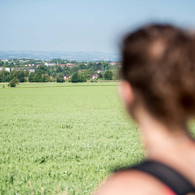 Frau mit Rucksack blickt auf eine grüne Wiese und eine weite Landschaft mit Häusern im Hintergrund.