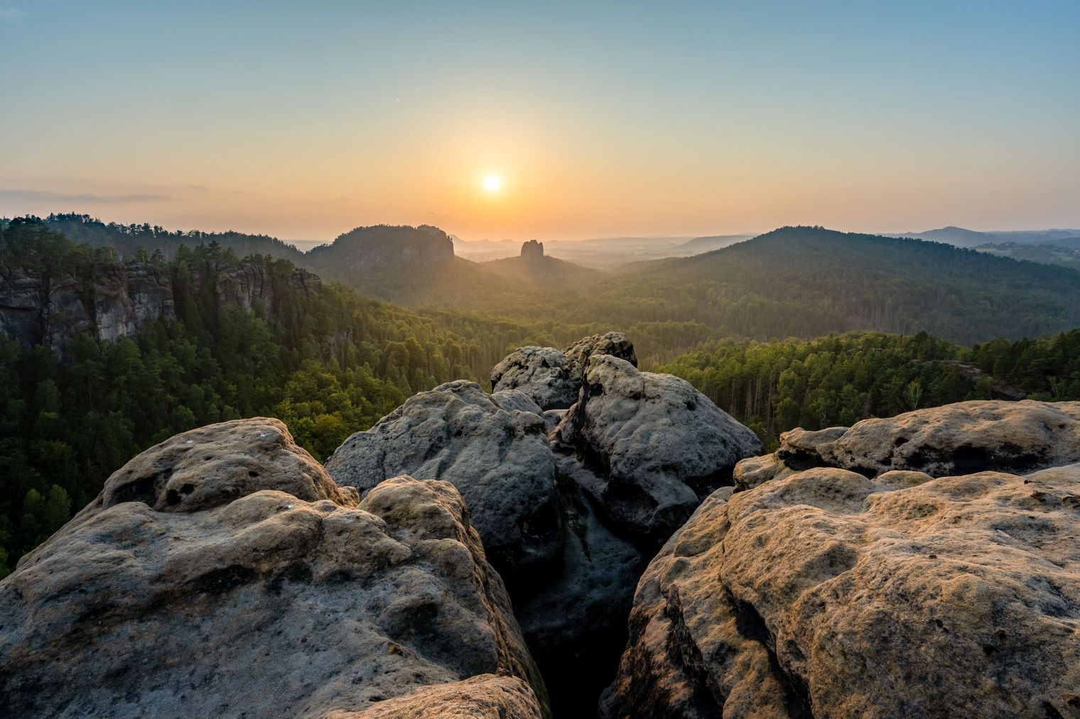 Sonnenaufgang über einer bewaldeten Landschaft mit Felsen im Vordergrund.