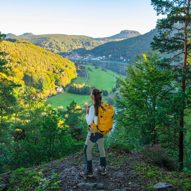 Wanderin mit gelbem Rucksack steht auf einem Hügel und blickt auf eine grüne Tallandschaft.