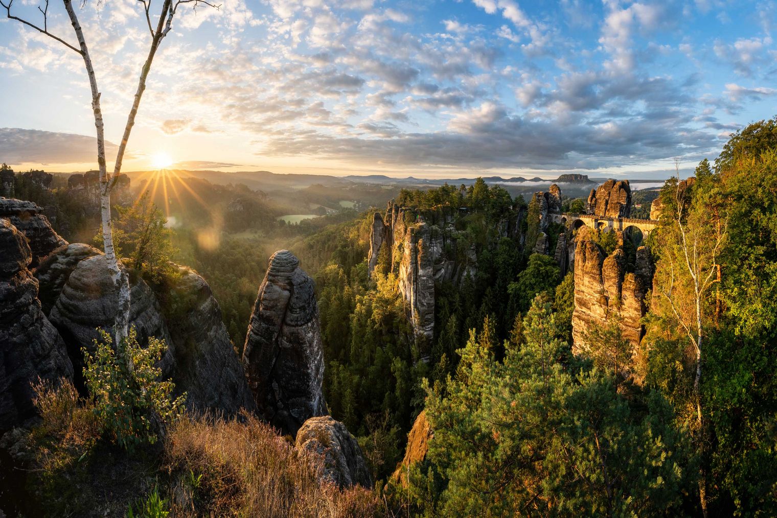 Sonnenaufgang über einer felsigen Landschaft mit Bäumen und Wolken am Himmel.