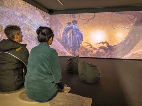 Zwei Personen sitzen auf Bank und schauen sich virtuelle Ausstellung zu Caspar David Friedrich an.