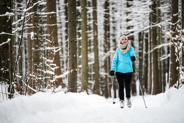 Frau in blauer Jacke wandert mit Skistöcken durch einen verschneiten Wald.