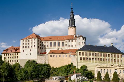Imposantes, historisches Schloss Weesenstein mit Turm, mit Bäumen am Fuße des Schlosses und blauem Himmel mit Wolken.