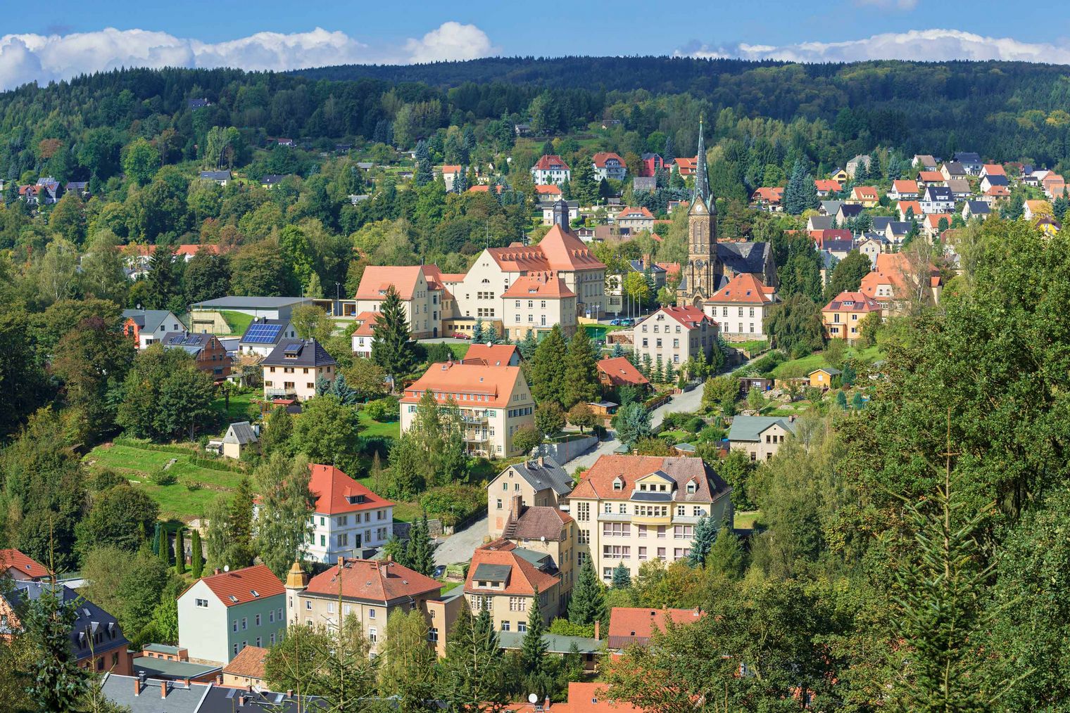 Blick auf eine malerische Stadt mit roten Dächern, umgeben von grünen Hügeln und Bäumen.