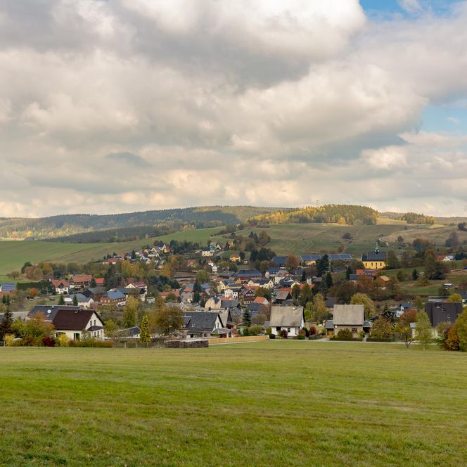 Blick auf eine kleine Stadt mit Häusern, umgeben von grünen Feldern und Hügeln unter einem bewölkten Himmel.