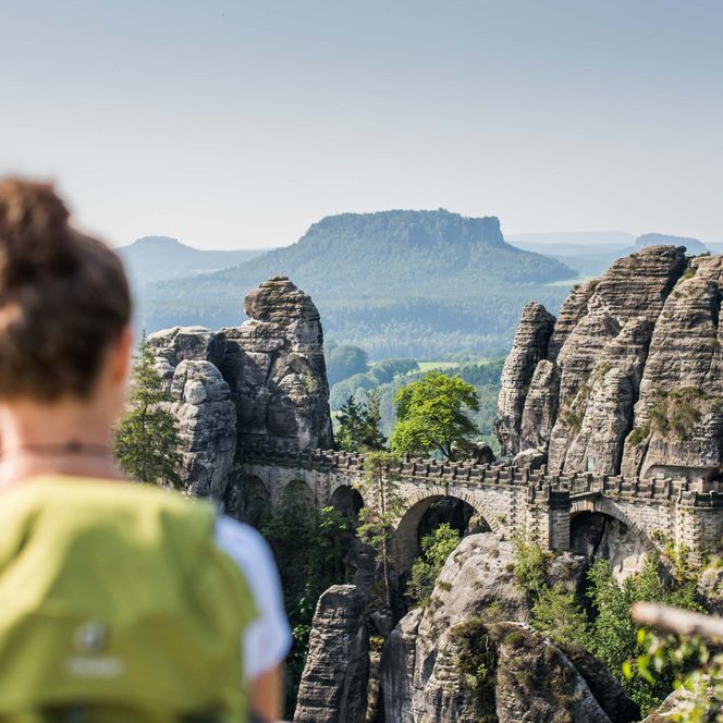 Frau mit Rucksack blickt auf die Felsen der Bastei und dem Lilienstein im Hintergrund.