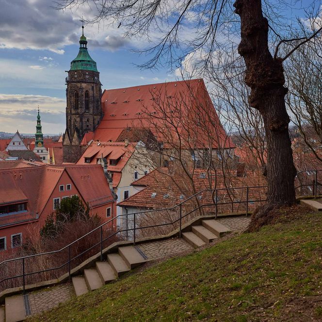 Blick auf eine Stadt mit roten Dächern und einer Kirche mit grünem Turm, umgeben von Bäumen.