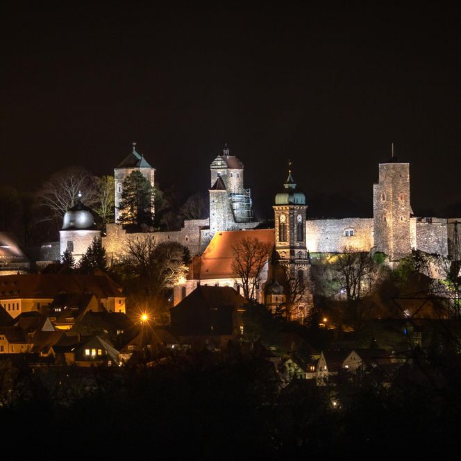 Beleuchtete Burganlage auf einem Hügel bei Nacht, umgeben von Bäumen und Stadtlichtern.