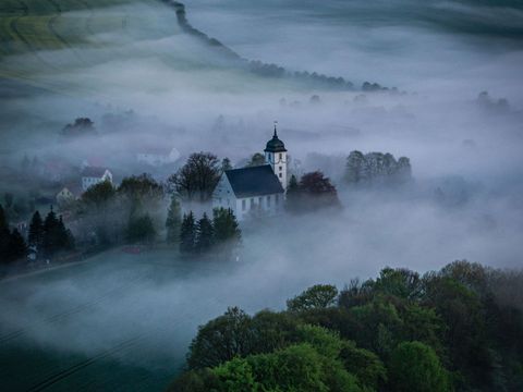 Kirche in Gohrisch mit weißem Turm und dunklem Dach, umgeben von Nebel und dunklem Wald..