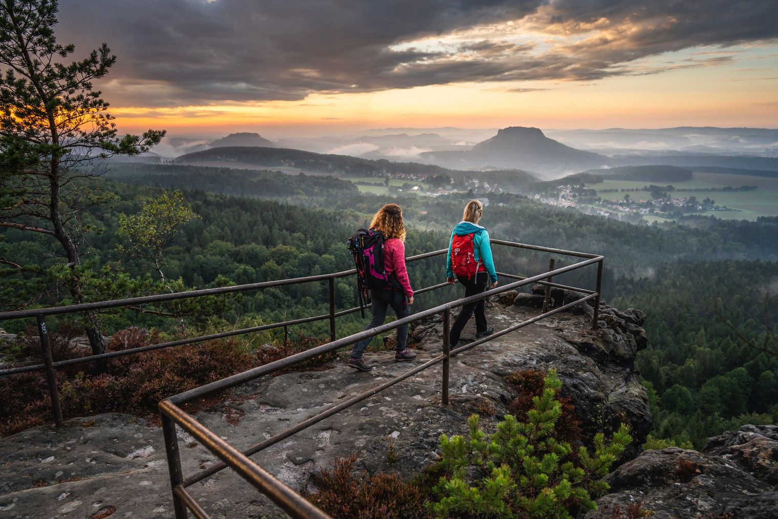 Zwei Wanderer mit Rucksäcken stehen an einem Aussichtspunkt mit Blick auf eine bewaldete Landschaft und einen Sonnena...