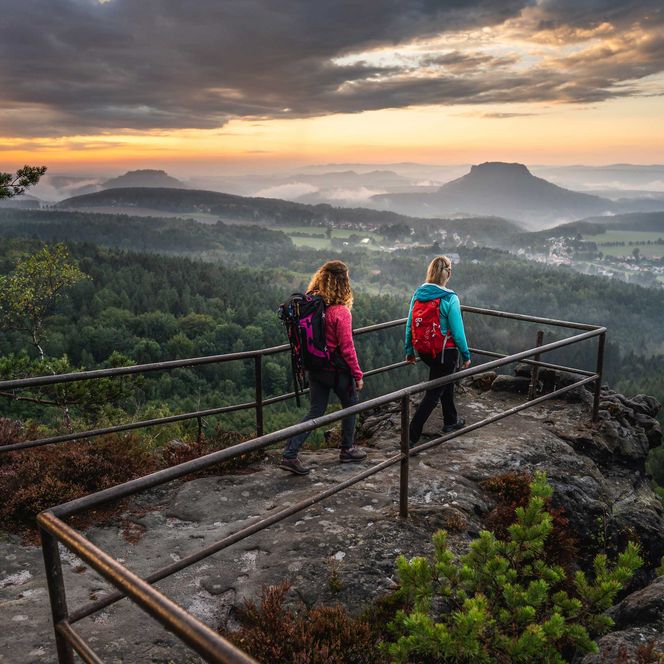Zwei Wanderer mit Rucksäcken stehen an einem Aussichtspunkt mit Blick auf eine bewaldete Landschaft und einen Sonnena...