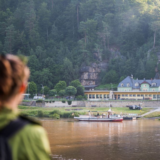 Wanderer mit Rucksack blickt auf ein Gebäude am Fluss, umgeben von grünen Felsen.
