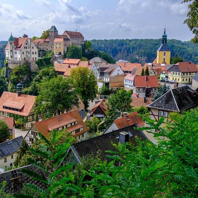 Blick auf eine malerische Stadt mit bunten Häusern und einer Burg im Hintergrund, umgeben von Bäumen.