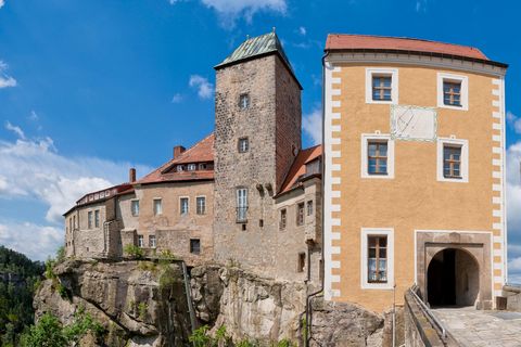 Ansicht des Einganges der Burg Hohnstein mit einem hohen Turm und orangefarbenen Wänden, umgeben von grünen Bäumen und blauem Himmel.