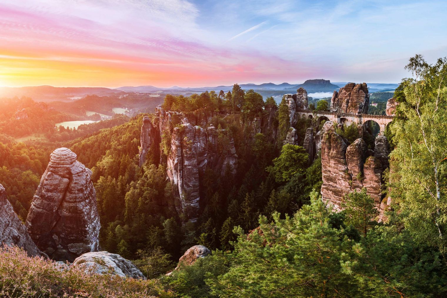 Sonnenuntergang über einer felsigen Landschaft mit Bäumen und Bergen im Hintergrund.