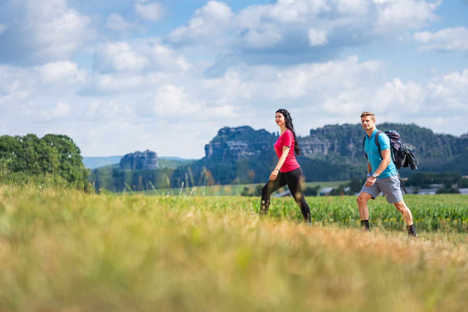 Zwei Wanderer gehen auf einem Feld, im Hintergrund sind Felsen und ein blauer Himmel mit Wolken.