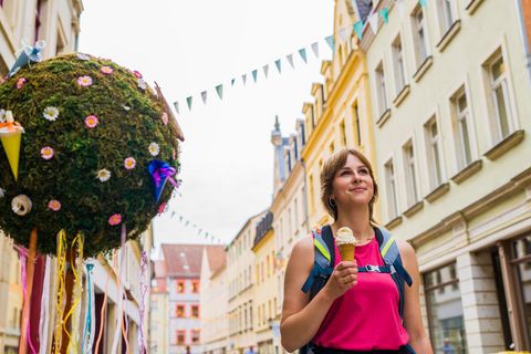 Frau mit Rucksack hält ein Eis in einer festlich geschmückten Straße mit bunten Fähnchen.