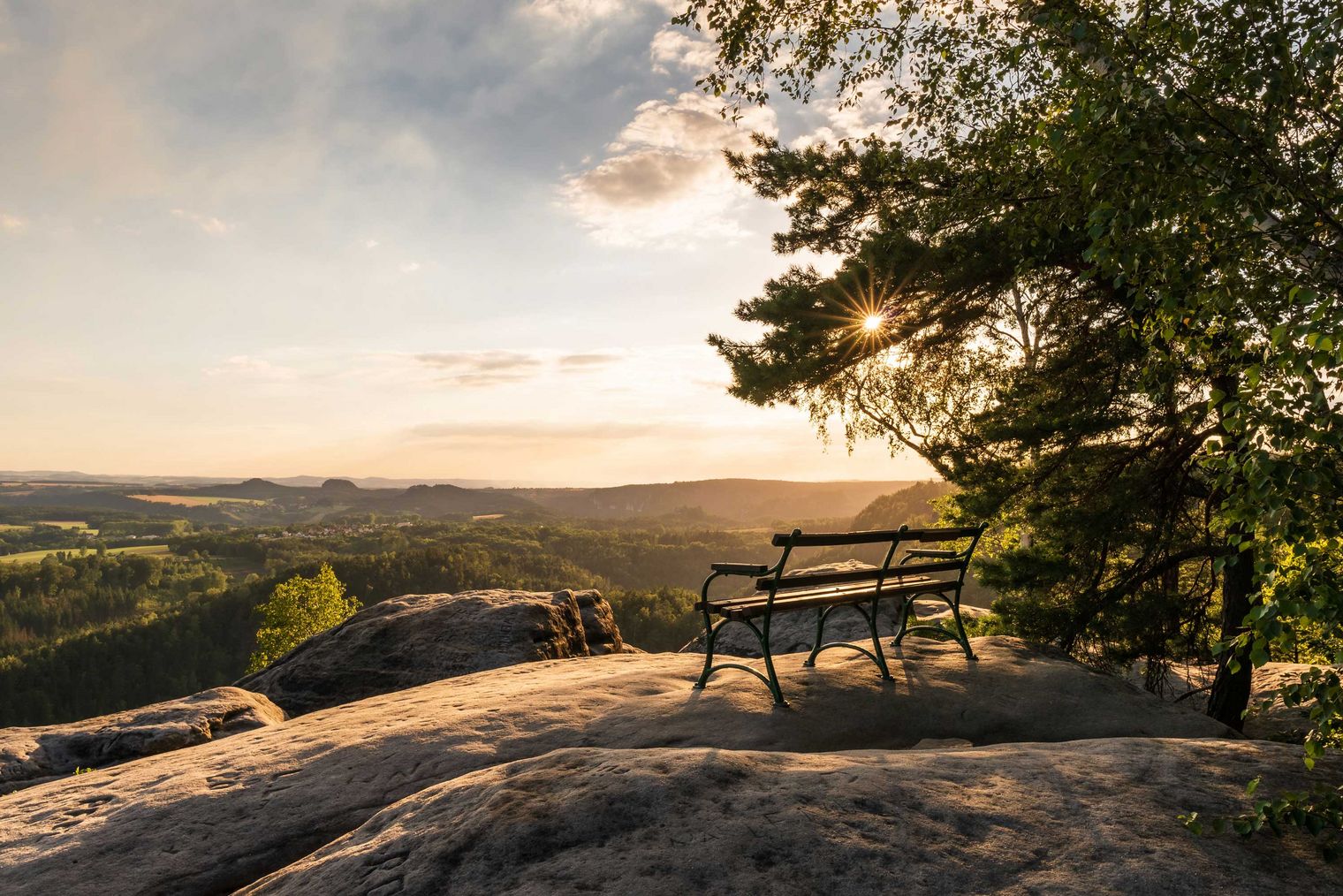 Blick auf die Landschaft mit einer Bank auf einem Felsen, umgeben von Bäumen bei Sonnenuntergang.
