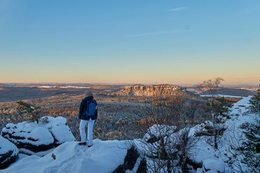 Person in blauer Jacke steht auf schneebedecktem Felsen und blickt auf eine winterliche Landschaft.