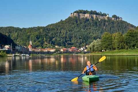Mann im Kajak paddelt auf ruhigem Wasser, im Hintergrund eine Stadt und ein bewaldeter Hügel mit einer Festung.