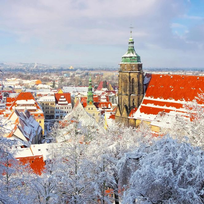 Schneebedeckte Dächer und eine Kirche mit grünem Turm unter einem blauen Himmel.