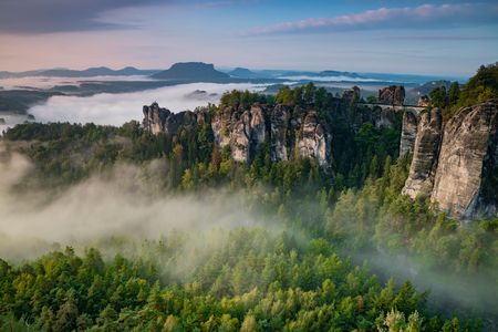 Waldige Felsformationen ragen aus Nebel, im Hintergrund Hügel bei Sonnenaufgang.