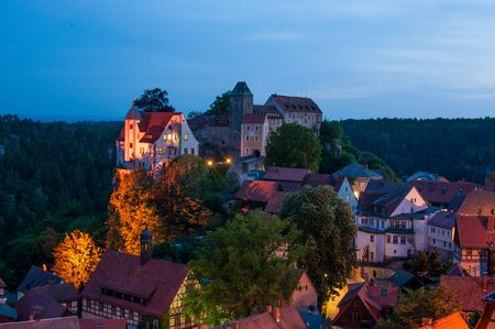 Blick auf eine Stadt mit historischen Gebäuden und einer Burg, umgeben von Bäumen und sanften Hügeln bei Dämmerung.